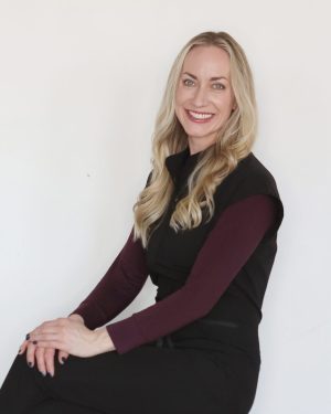 Smiling woman with long blonde hair, wearing a black and burgundy outfit, sitting against a white background. Smiling woman with long blonde hair, wearing a black and burgundy outfit, sitting against a white background.